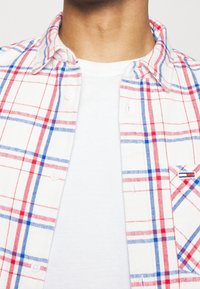 Närbild av en person som bär en vit t-shirt under en uppknäppt vit rut skjorta med röda och blå linjer.