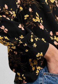Black floral blouse featuring yellow and pink flowers, long sleeves with gathered cuffs, and a relaxed fit, paired with denim jeans.