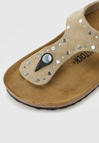 Beige suède teensandalen met metalen studs, zwart hardwareaccent en een gestructureerde bruine voetbed. De buitenzool heeft een gebogen ontwerp.