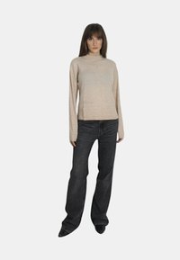 Maglione a maglia beige con collo alto, maniche lunghe e due tasche frontali, abbinato a jeans grigi scuri a gamba larga. Testura morbida, design casual.