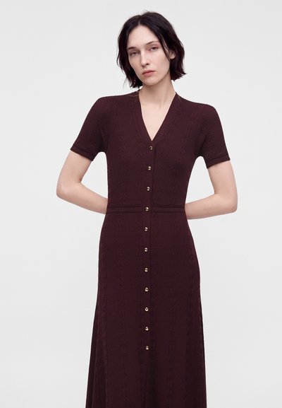 Femme aux cheveux courts et foncés portant une robe marron foncé ajustée, à manches courtes, boutonnée, avec des motifs texturés subtils sur un fond uni.
