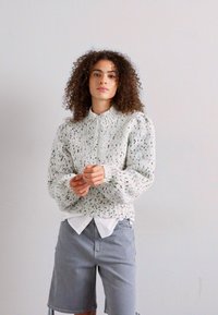 Strickpullover mit hohem Kragen in hellgrau, mit einem strukturierten Muster und puffärmel. Kombiniert mit grauen Shorts und einem weißen Shirt.