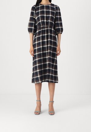 Kobieta ubrana w midi sukienkę w kratę w kolorach granatowym, białym i brązowym, z rękawami o długości trzy czwarte typu bufka oraz w jasnoniebieskie sandały na wysokim obcasie z paskiem wokół kostki.