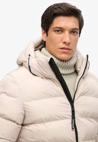 Chaqueta acolchada beige con capucha y cremallera negra, con paneles acolchados horizontales. Lleva puesta una sweatshirt de punto gris claro con cuello alto.