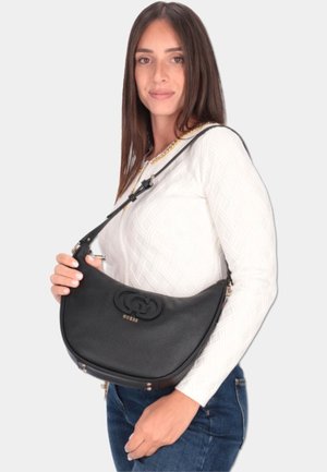 Borsa a tracolla in pelle nera dalla forma curva, con logo in rilievo e dettagli in metallo dorato. Indossata con una maglia bianca a fantasia e un paio di jeans casual.