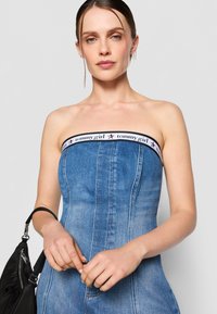 Denim strapless dress with a fitted silhouette, featuring a black and white branded band at the neckline. Styled with a black handbag.
