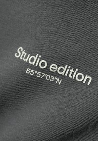 Grijze stof met een gestructureerd oppervlak heeft witte borduursels met de tekst "Studio editie" en "55°57'03"N" die prominent worden weergegeven.