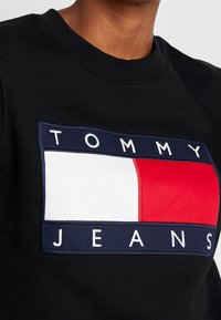 Mustapaita, jossa on suuri suorakaiteen muotoinen Tommy Jeans -logo punaisen, valkoisen ja sinisen värimaailman kanssa. Ribattua kaula-aukkoa ja teksturoitua kangasta.