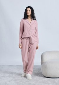 Γυναίκα που στέκεται με ροζ και λευκή καρό πιτζάμα και λευκές παντόφλες σε γκρι χαλί δίπλα σε εκρού καμπυλωτό καναπέ, ουδέτερο φόντο.