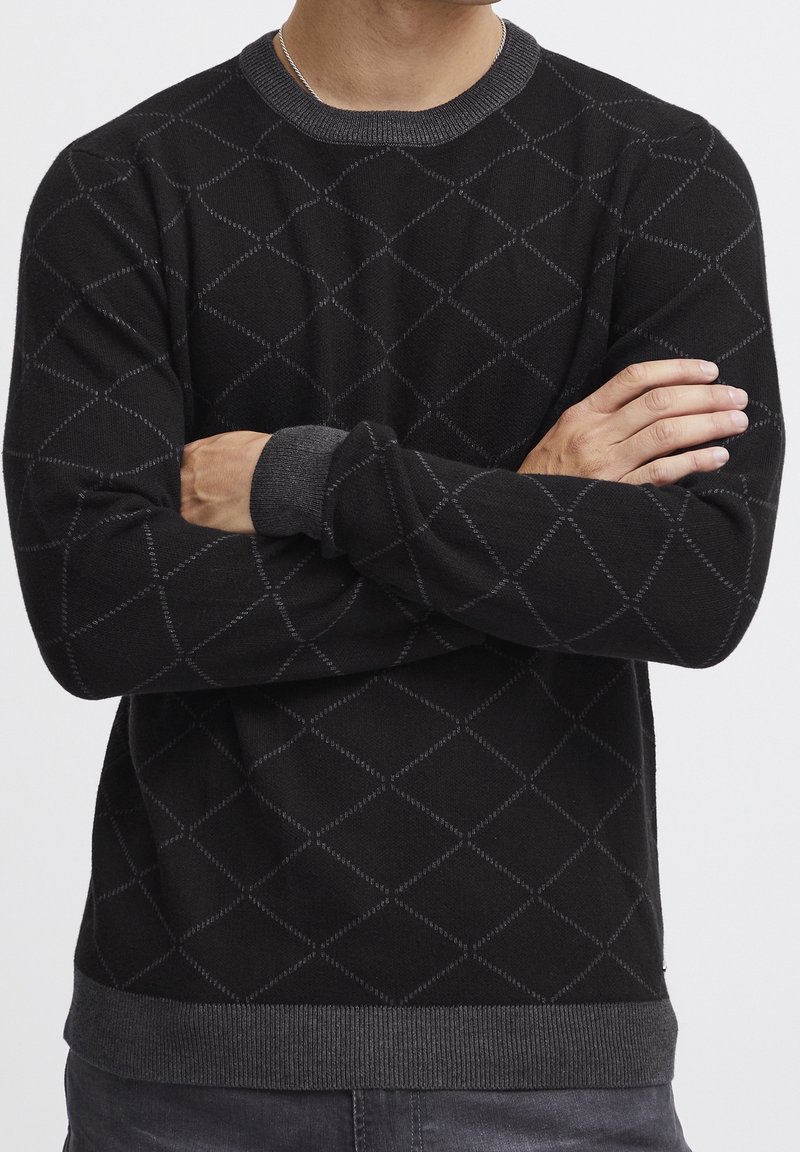 Zwarte trui met een diamanten patroon, geribde halslijn en manchet, met een zachte textuur. Het ontwerp heeft contrasterende grijze accenten.