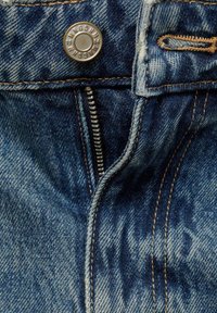 Gros plan sur un jean en denim bleu foncé montrant une braguette partiellement zippée avec un bouton argenté et des détails de coutures orange.