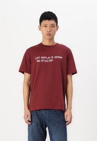Červené tričko s krátkým rukávem s bílým nápisem "LOT REPLAY B. DENIM RD 39 423 925" umístěným ve středu na přední straně, vyrobené z jemné bavlněné tkaniny.