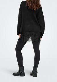 Femme debout de dos, portant un pull en maille noir sur un top en dentelle noire, des leggings noirs et des bottines noires.