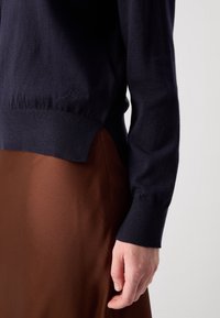 Pull à manches longues bleu marine avec détail fendu, porté sur une jupe marron texturée, montrant le bras et la main gauches sur un fond uni.