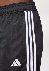 Black sports shorts made of smooth fabric, featuring three white vertical stripes and a white logo on the lower left side.