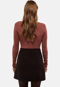 Femme aux longs cheveux bruns ondulés, portant un pull côtelé rose poudré et une jupe taille haute brun foncé, tournée de dos.