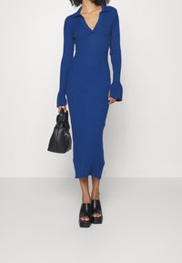 Abito blu a maglia rigata con colletto, maniche lunghe e polsini svasati. Abbinato a una borsa nera e sandali neri con plateau spessi.