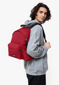 Sac à dos rouge fabriqué en tissu durable, avec un dessus arrondi, une poche avant, des zips noirs et des bretelles réglables noires. Design minimaliste.