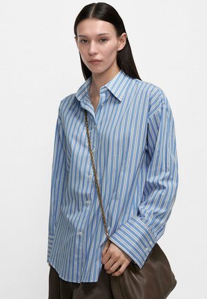 Kobieta o długich ciemnych włosach, ubrana w niebiesko-białą koszulę w paski z guzikami, trzymająca brązową skórzaną torbę z złotym łańcuszkiem.