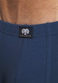 Boxers bleus en coton avec une étiquette noire portant un logo de bélier. Le tissu a une texture lisse et un design simple avec des détails de couture.