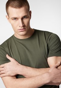 Zielona bawełniana koszulka z okrągłym dekoltem i krótkimi rękawami, o miękkiej teksturze i dopasowanym kroju, noszona przez męskiego modela.