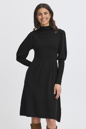 Femme souriante portant une robe noire à manches longues avec des épaules bouffantes et des bottes marron remontant jusqu’aux genoux sur un fond uni.