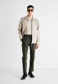 Veste zippée beige clair sur un T-shirt beige, assortie à un pantalon tailleur vert foncé et des chaussures noires. Des lunettes de soleil rondes complètent le look.