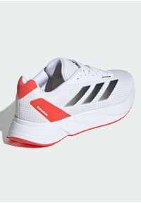 Zapatilla deportiva blanca con parte superior de malla, rayas negras y un talón y acentos rojos texturizados. Cuenta con una suela acolchada y un diseño ligero.