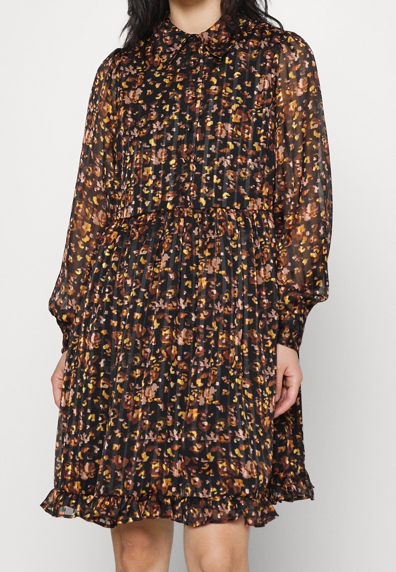 Femme portant une robe noire à manches longues avec un motif floral multicolore, jupe plissée et ourlet à volants, debout devant un fond uni.