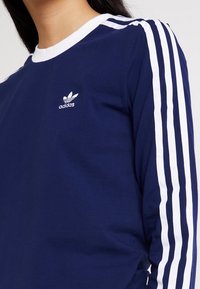 Laivastonsininen pitkähihainen paita, joka on tehty puuvillasta. Paidassa on valkoiset kolmiomerkinnät hihoissa ja pieni valkoinen Adidas-logo rinnassa.