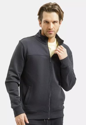 Mężczyzna w ciemnoszarej kurtce zapinanej na zamek na jasną koszulę, poprawiający kołnierz prawą ręką, zwrócony przodem na białym tle.