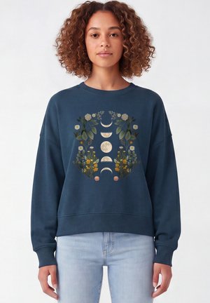 Jeune femme aux cheveux bouclés porte un sweat-shirt bleu avec des phases lunaires et des motifs floraux sur le devant, associé à un jean bleu clair.