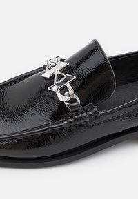 Mocassin en cuir noir brillant avec une chaîne en métal argenté et une boucle décorative abstraite sur le devant supérieur.