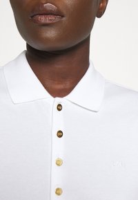 Vit polo skjorta med krage, prydd med knappar i guldton och en liten broderad logotyp. Texturerad tyg med klassisk passform.