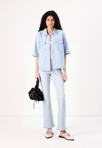 Jaqueta de ganga azul clara com mangas curtas, dois bolsos frontais, combinada com jeans flare azuis claros e uma mochila preta com estampado floral e cordão.
