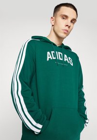 Grön hoodie med känguruficka, dragsko i huvan och vitt Adidas-logotyp med kontrasterande vita ränder längs ärmarna.