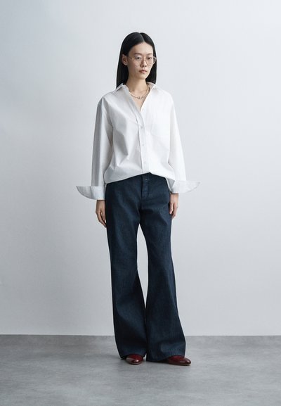 Femme aux cheveux noirs et raides portant des lunettes, une chemise blanche oversize, un pantalon large foncé et des chaussures bordeaux, debout contre un mur gris uni.
