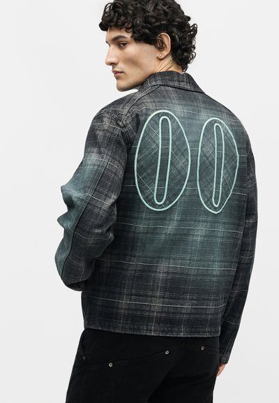 Άνδρας μοντέλο με σγουρά μαλλιά που φοράει ένα σκούρο πράσινο καρό μπουφάν με μεγάλο ραμμένο διπλό μηδέν "00" στην πλάτη.