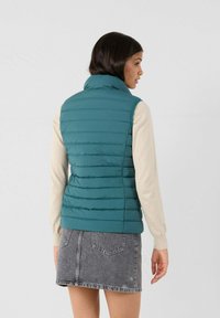 Gilet imbottito verde acqua con colletto alto e cuciture orizzontali, indossato sopra un maglione leggero. Abbinato a una gonna di jeans grigia.