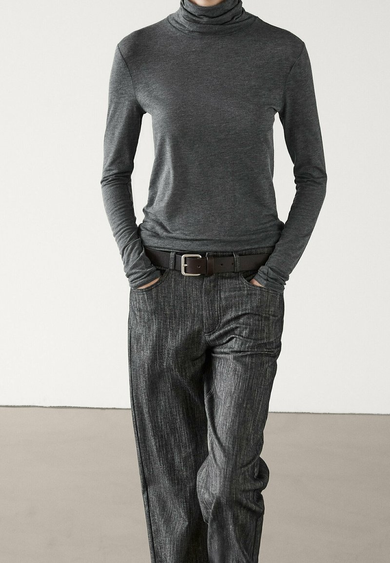 Personne portant un col roulé gris foncé ajusté et un pantalon texturé gris foncé ample, les mains dans les poches et une ceinture noire.