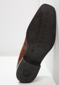 Chaussure en cuir marron avec une semelle en caoutchouc noire. La semelle présente des détails texturés et un logo. La forme est élégante avec des bords incurvés.