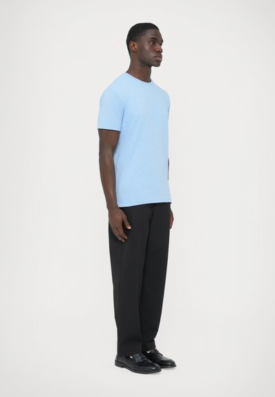 Γαλάζια κοντομάνικη μπλούζα από βαμβάκι, συνδυασμένη με σκούρο παντελόνι και μαύρα παπούτσια, παρουσιάζεται σε πλευρική όψη με ουδέτερο φόντο.