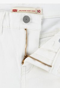 Bijele traperice s kratkim nogavicama, sa zlatnim patentnim zatvaračem, metalnim dugmetom i oznakom koja pokazuje "Levi's Girlfriend Shorty Short" u veličini 10.
