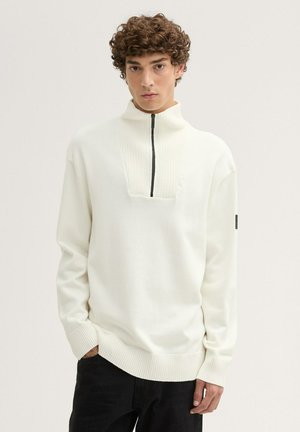 Joven con cabello rizado que lleva un suéter blanco con media cremallera y pantalones negros, de pie con una mano en el bolsillo frente a un fondo liso.