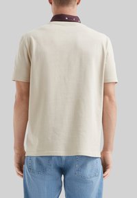 T-shirt beige con maniche corte e colletto bordeaux caratterizzato da un motivo sottile. Materiale morbido e vestibilità rilassata. Vista posteriore mostrata.