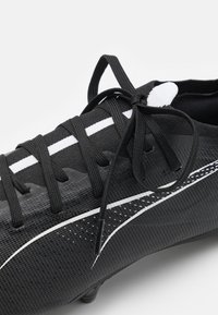 Chaussure de football noire avec une tige texturée, des lignes d'accentuation blanches et des lacets noirs. Détails notables incluant un motif distinct sur le matériau.