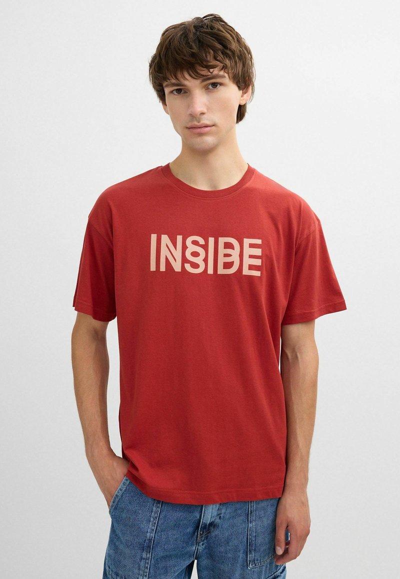 Joven con cabello castaño que lleva una camiseta roja con la palabra "INSIDE" en letras beige, combinada con jeans de mezclilla azul.