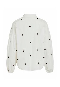 Blouse blanche manches longues avec des motifs de cœurs noirs dispersés sur le tissu, dotée d’un col et de manches bouffantes avec poignets élastiques.