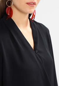 Femme portant un chemisier noir à col en V et de grandes boucles d'oreilles créoles dorées avec des pendentifs translucides rouges sur fond blanc.