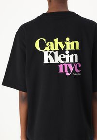Juodos medvilnės marškinėliai su dideliu, daugiaspalviu atspaudiniu: "Calvin Klein" gelsvu ir baltu, "nyc" rožiniu, su maža logotipu po juo.